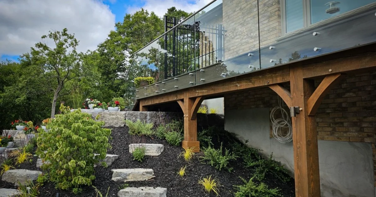Elevated deck with glass railing supported by timber posts, surrounded by lush landscaping and stone steps.